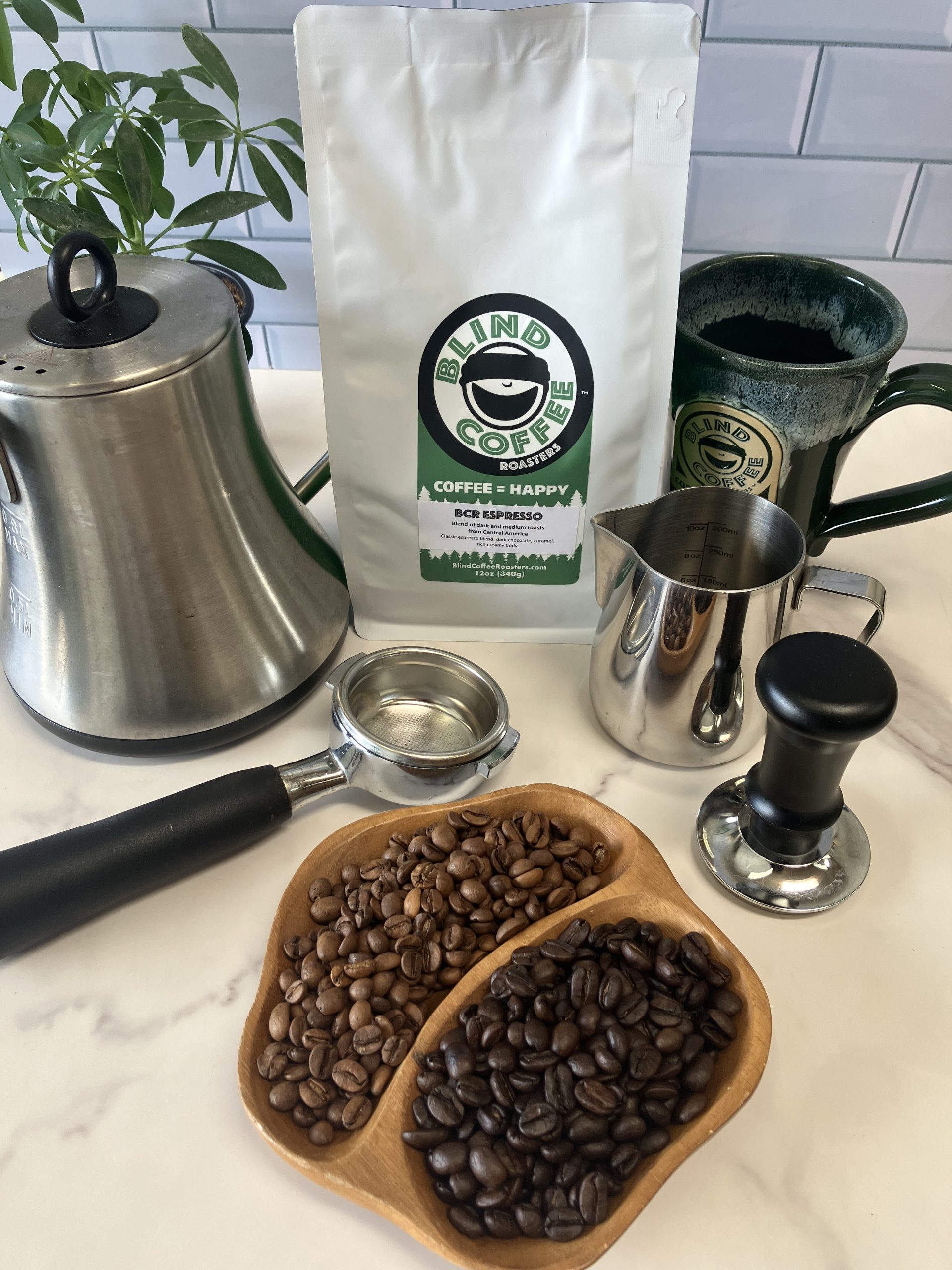 A 12oz bag of BCR Espresso on a kitchen counter with a gooseneck kettle, mug, milk pitcher, tamper, portafilter and divided bowl filled with both roasted coffee beans of the blend.