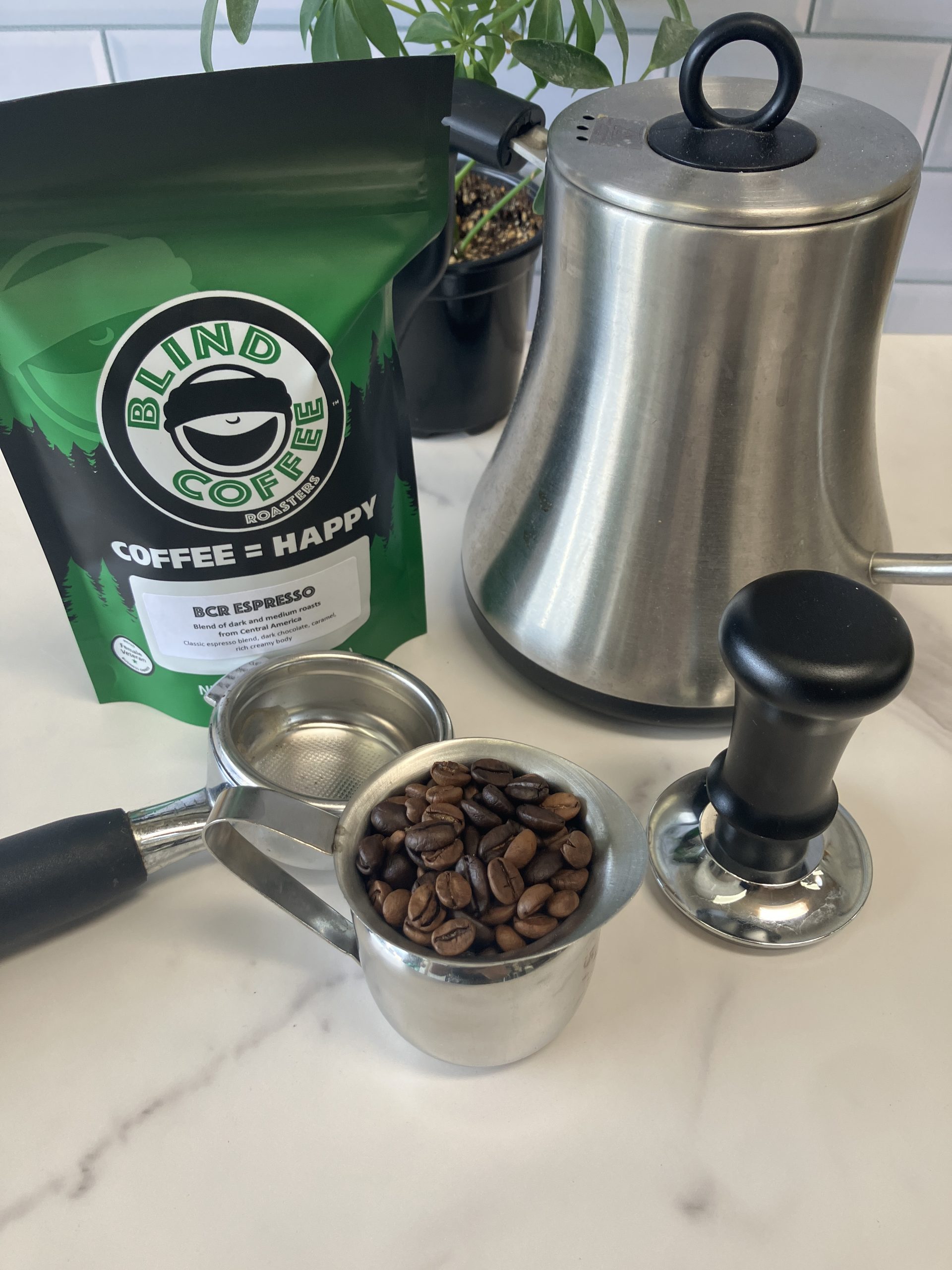 A 6oz bag of BCR Espresso on a kitchen counter with a gooseneck kettle, portafilter, tamper, portafilter and espresso cup filled with both roasted coffee beans of the blend.