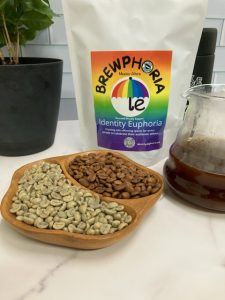 Photo showing close up of green and roasted Brewphoria beans with bag of Brewphoria, pour over pitcher, coffee canister, and small coffee tree.