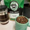 Photo showing close up of roasted Guatemalan beans with a bag of ER's Favorite Guatemala, coffee press, coffee canister, and small coffee tree in the background.