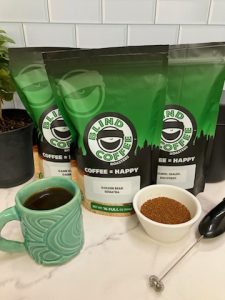 Photo showing 3 bags of coffee with bowl of ground coffee, milk frother, mug, and small coffee tree.