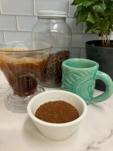 Kitchen counter with a small bowl full of coffee grounds, mug, pour over brewer, coffee canister, and small coffee tree.
