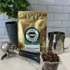 Photo showing 8oz bag of Gateau a la Cannelle with roasted beans in an espresso glass, portafilter, milk pitcher, and small coffee tree.