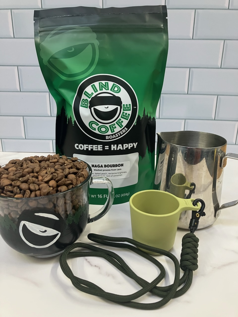 Naga Bourbon coffee beans in a glass cup with a bag of Naga Bourbon, milk steaming pitcher, and tasting cup.