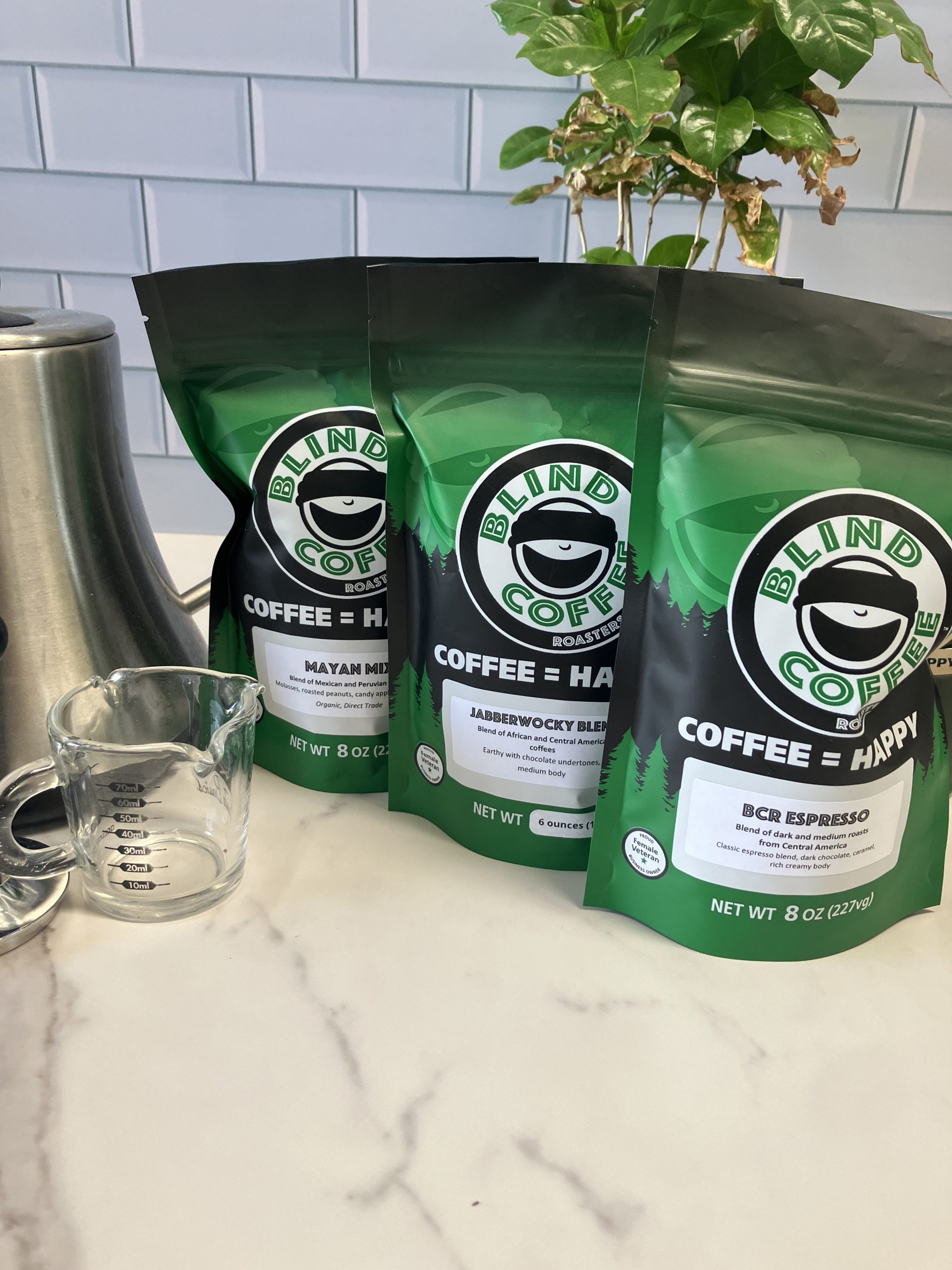 3 6oz bags of coffee on a kitchen counter with a gooseneck kettle, espresso, and coffee plant.