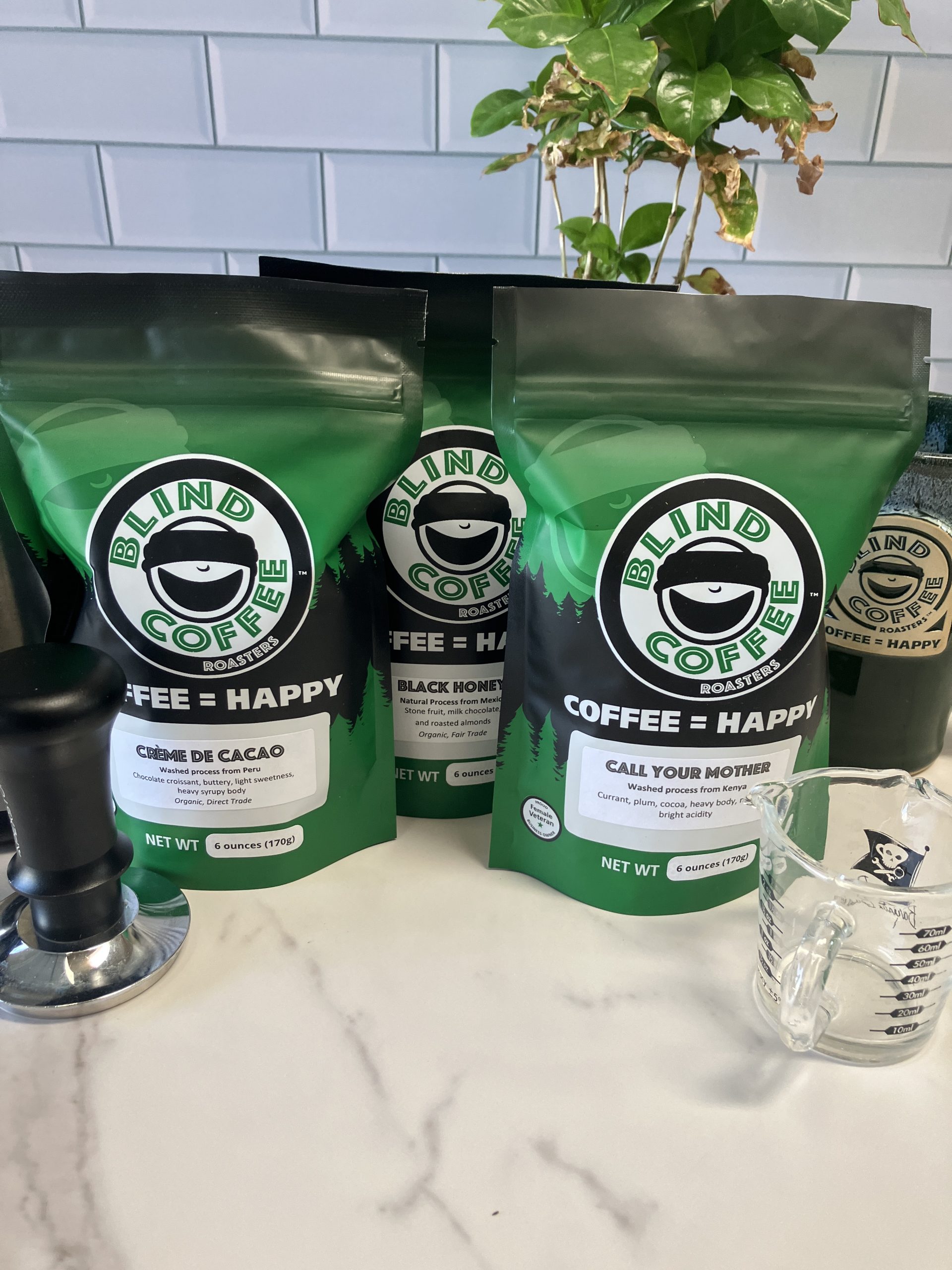 3 6oz bags of coffee on a kitchen counter with a gooseneck kettle, espresso, and coffee plant.
