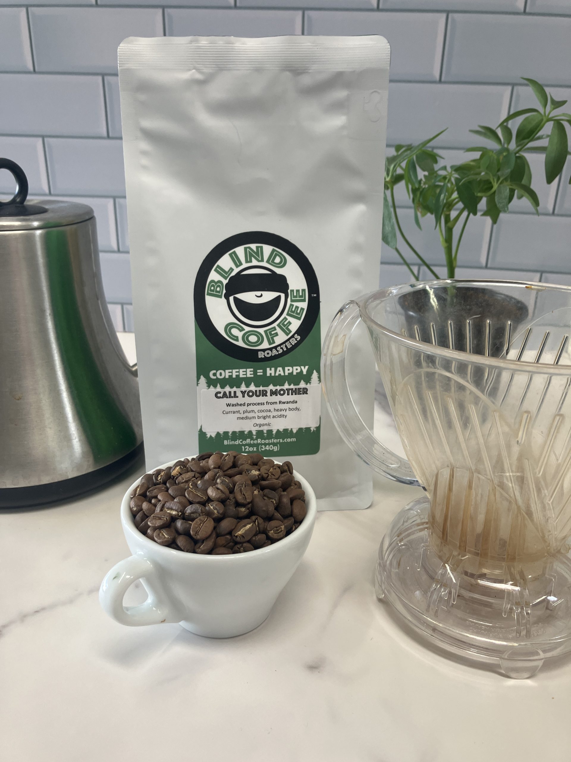 A 12oz bag of Call Your Mother on a kitchen counter with a gooseneck kettle, pour over device, and demitasse cup filled with roasted coffee beans.