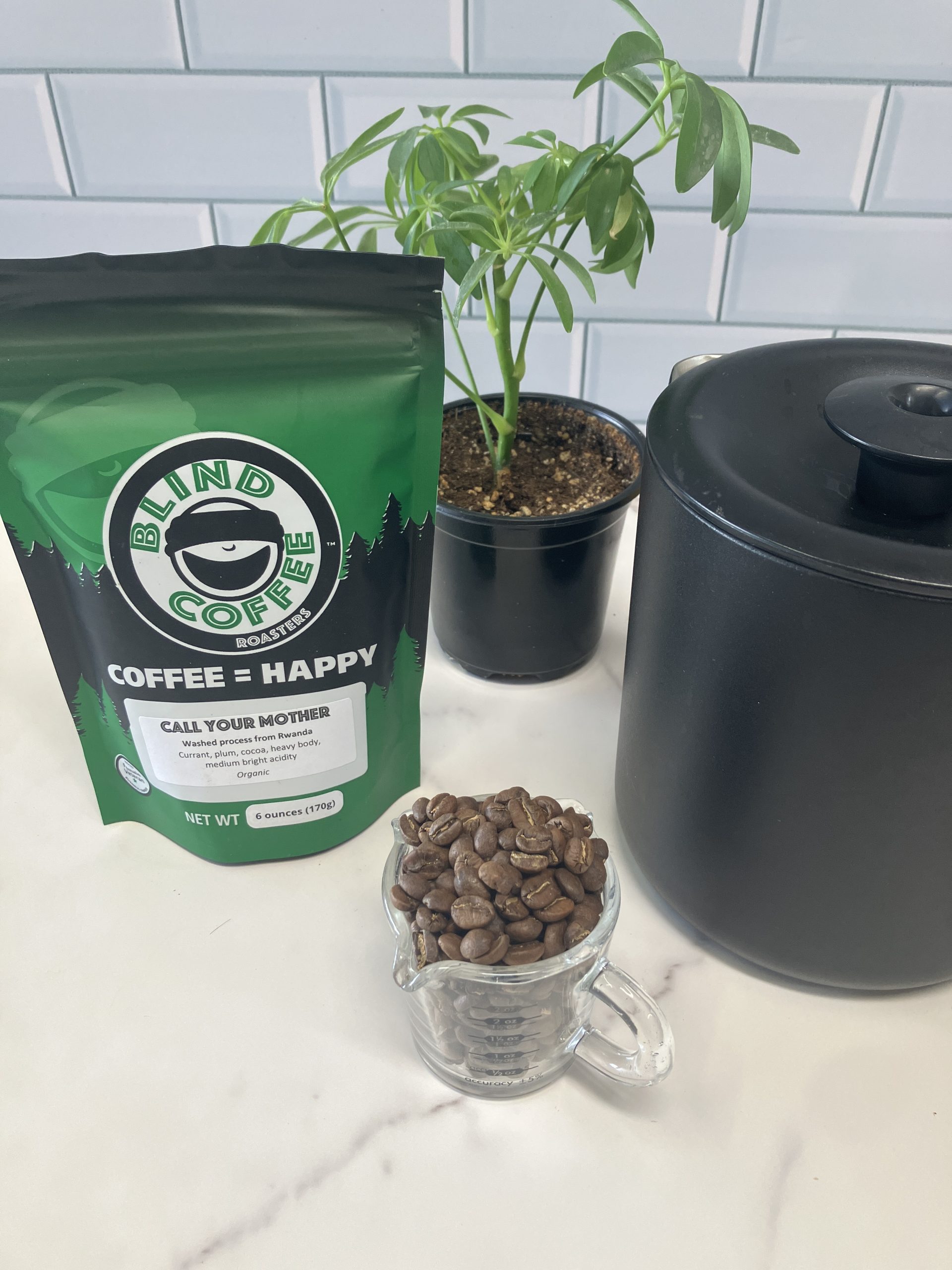 A 6oz bag of Call Your Mother on a kitchen counter with a coffee carafe and espresso pitcher filled with roasted coffee beans.