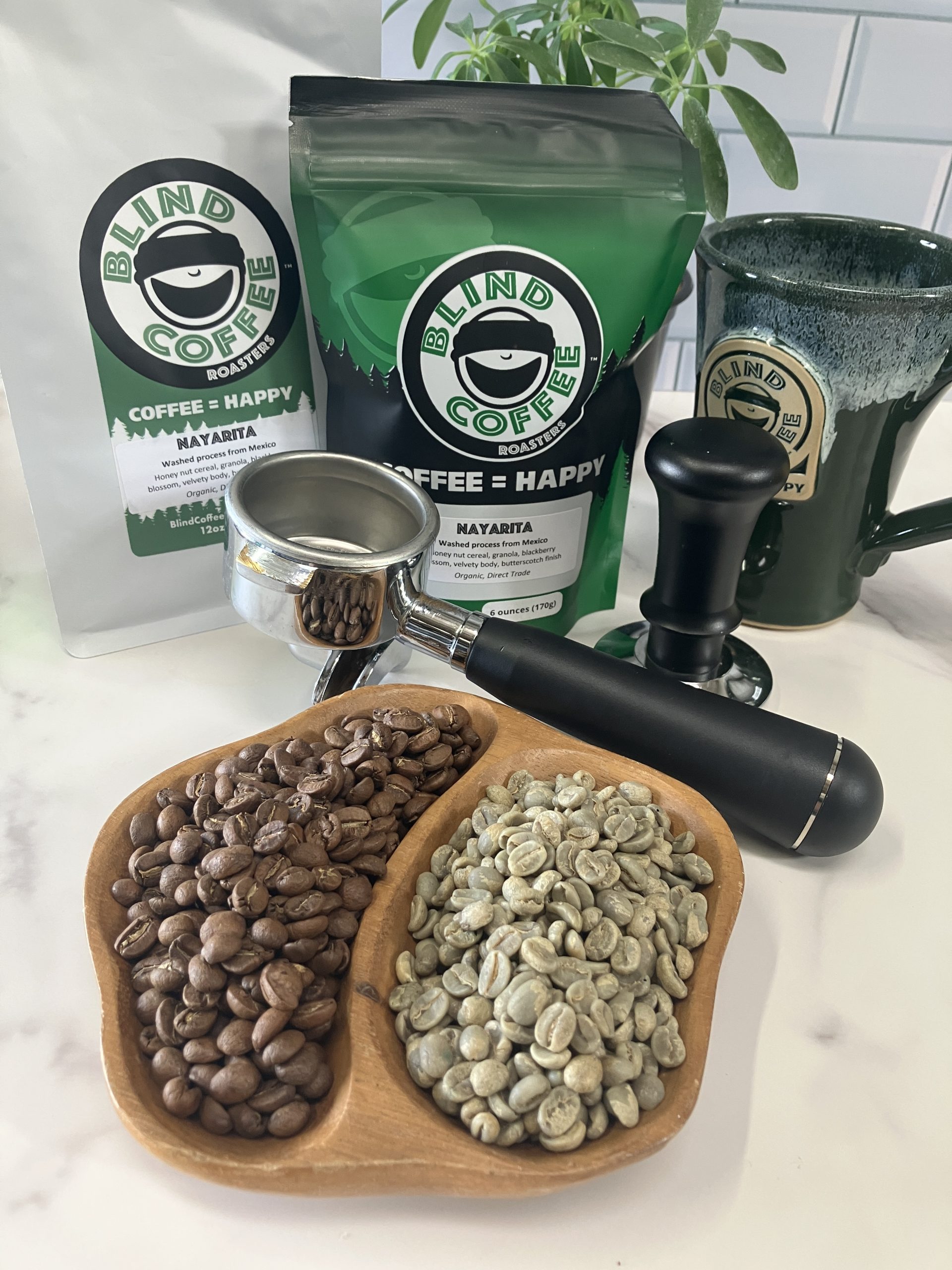 2 bags of Nayarita on a kitchen counter with a portafilter, tamper, white demitasse cup, and bowl filled with green & roasted coffee beans.